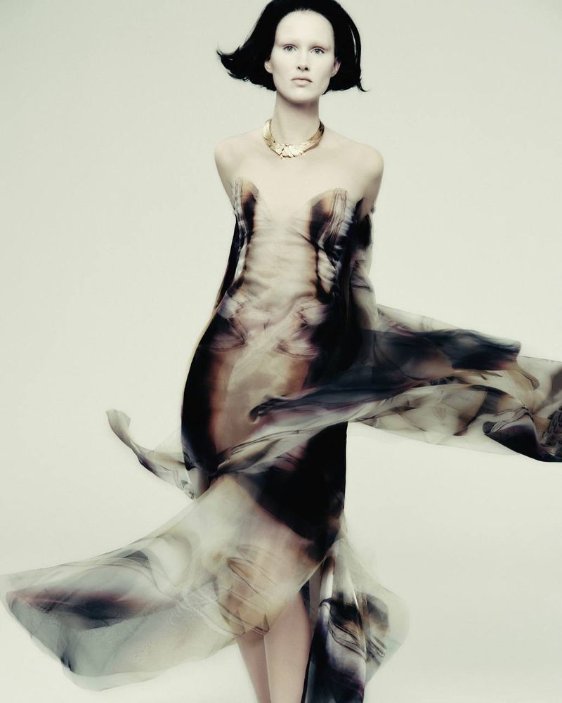 A slender adult female model stands in a photo studio wearing a flowing, semi-transparent dress, accessorized with a gold necklace against a neutral backdrop.