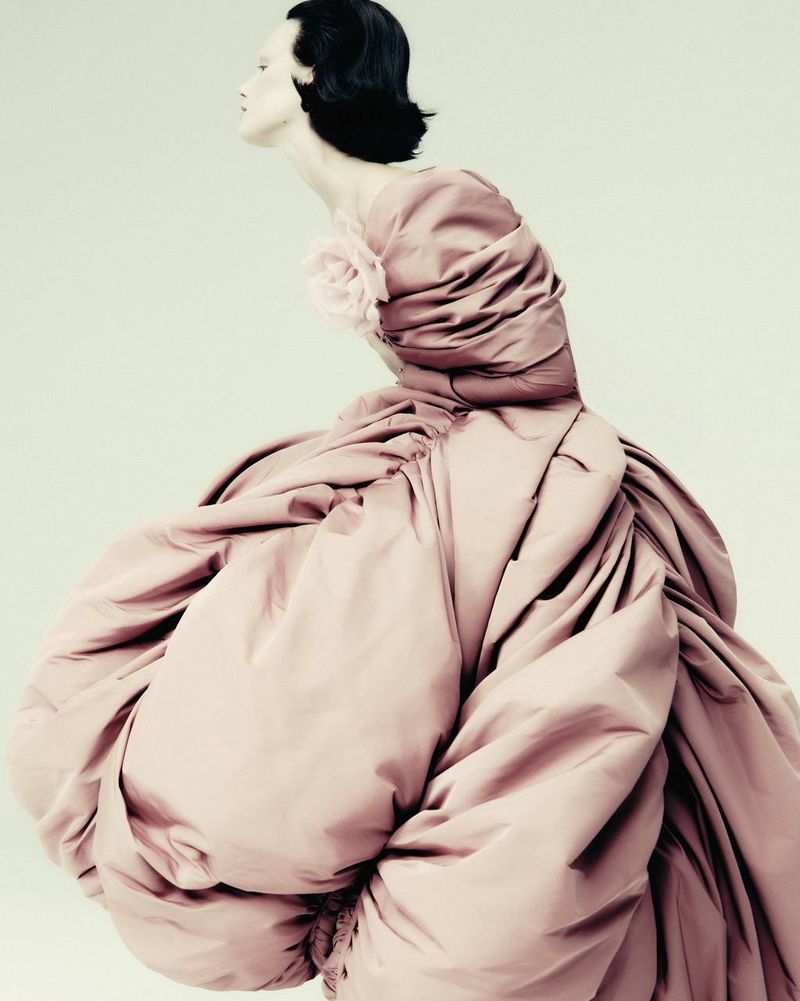 A side-view portrait of a model in a voluminous, gathered pink gown with a large floral detail, set against a clean, light background.