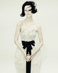 Model with dark short hair wearing an embellished strapless white gown with a black sash against a plain background