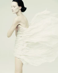 Fashion model in a strapless textured white dress with fabric billowing behind her against a pale studio backdrop.