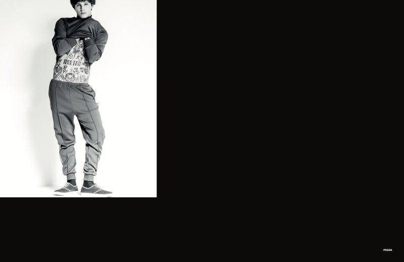 Young male model lifting dark sweatshirt to reveal tattooed torso against a stark white studio backdrop.