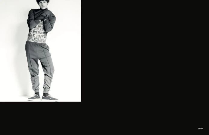 Young male model lifting dark sweatshirt to reveal tattooed torso against a stark white studio backdrop.