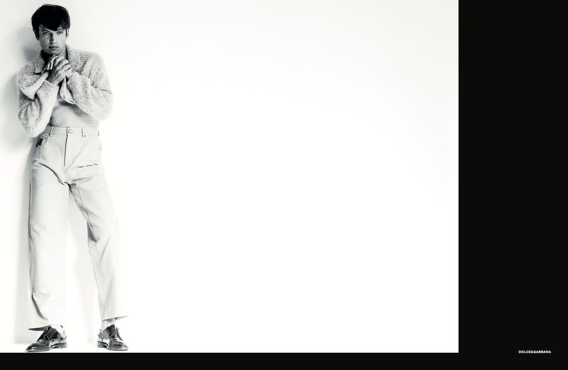 Model wearing a textured knit sweater and light trousers leaning against a white studio wall with black framing bars