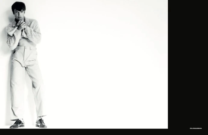 Model wearing a textured knit sweater and light trousers leaning against a white studio wall with black framing bars