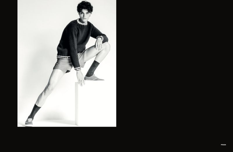 Model crouching on white pedestal wearing dark knit sweater and shorts for Numéro Homem Brasil editorial