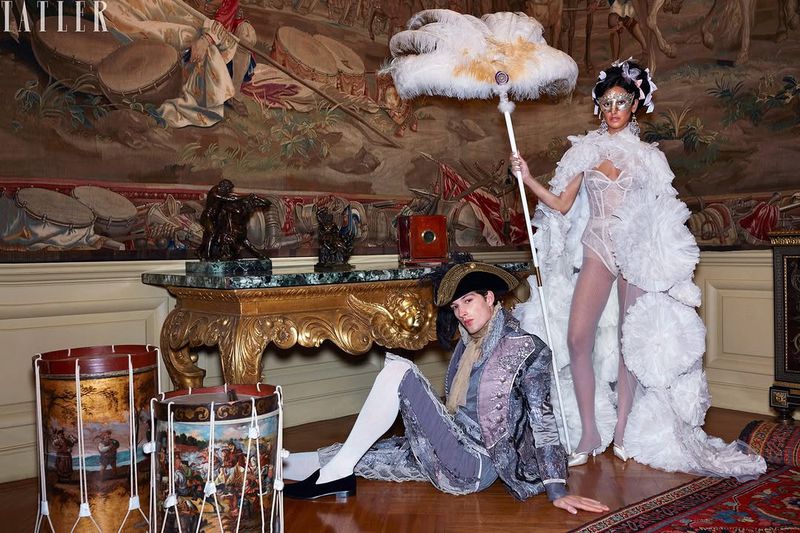 Two people dressed in period costumes in an ornate room. A tapestry hangs on the wall in the background. The image features unique props.