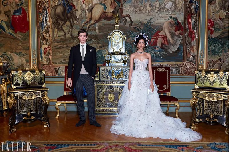 Man in a formal morning suit and woman in an elaborate white strapless wedding gown posing in an ornate room with a large wall tapestry.