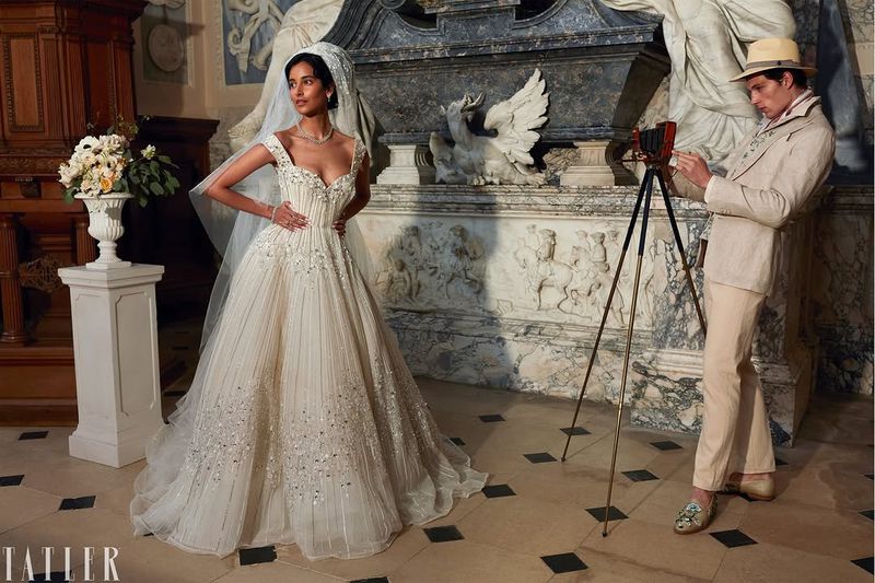 Model in embellished wedding gown and veil posing while a man in a linen suit operates a vintage camera on a tripod