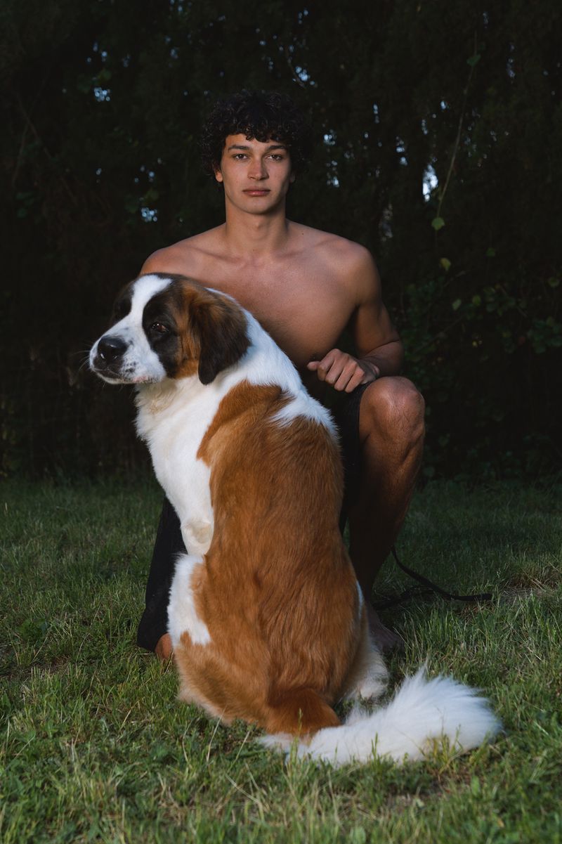 A shirtless male model kneeling outdoors on grass beside a large Saint Bernard dog against a dark foliage background.