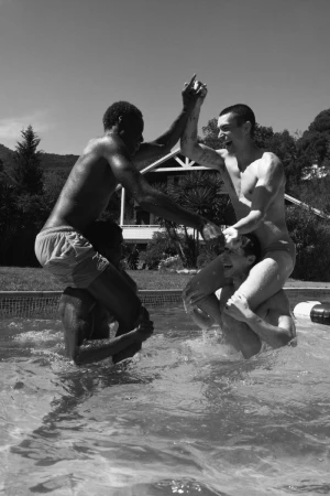 Four men playing chicken in a swimming pool, laughing and holding hands under clear daylight.