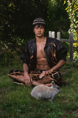 A man wearing a patterned jacket and cap sits cross-legged on grass next to a small rabbit, with foliage behind him.