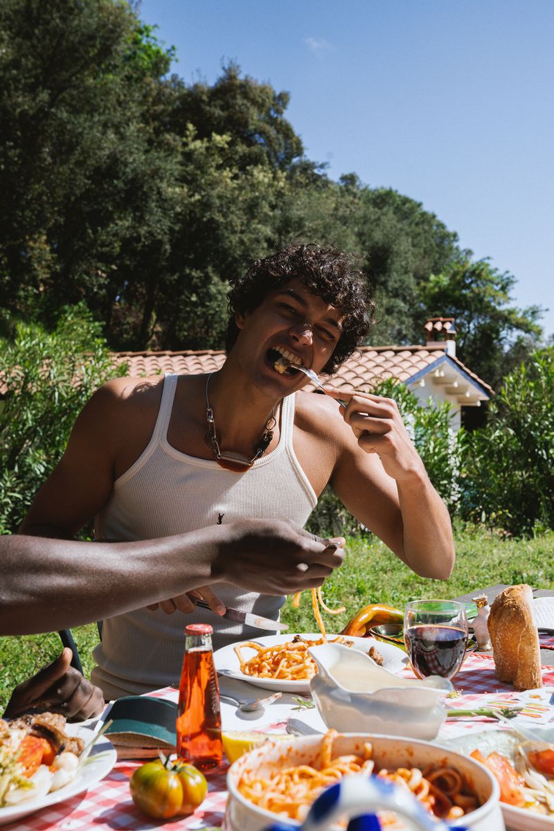 Man wearing a white tank top eating pasta outdoors at a table spread with food, drinks, and lush greenery in the background.
