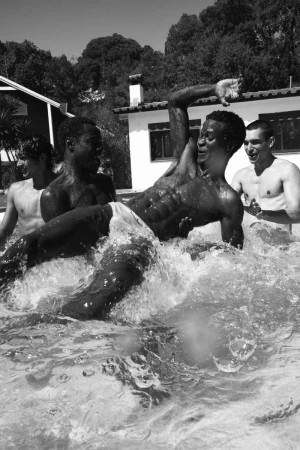 Four young men splashing and laughing in an outdoor swimming pool during the AS SUMMER COMES editorial project
