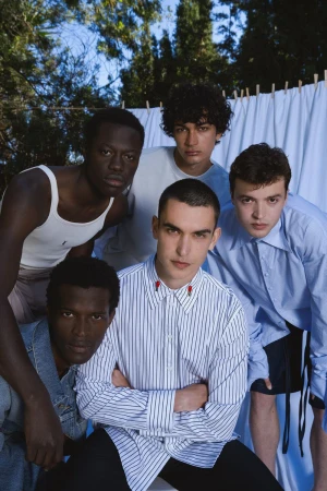 Group of five male models posing outdoors in front of a white fabric backdrop with trees visible above.