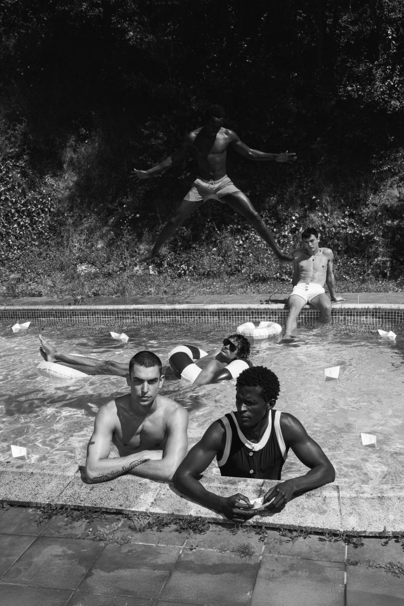Black and white editorial photograph of four male models in and around an outdoor swimming pool during summer