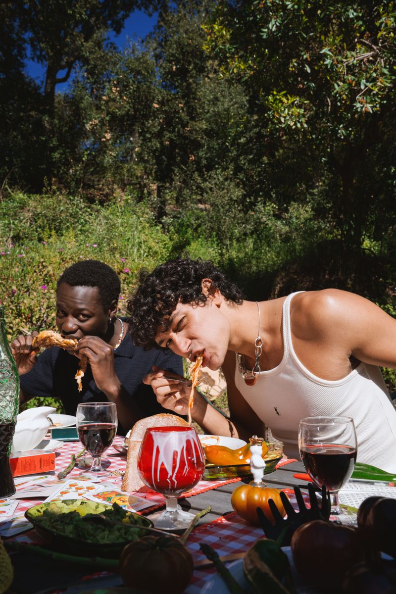 Two young men dining outdoors at a rustic table laden with food, wine glasses, and a red-patterned tablecloth under trees.