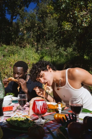 Two young men dining outdoors at a rustic table laden with food, wine glasses, and a red-patterned tablecloth under trees.