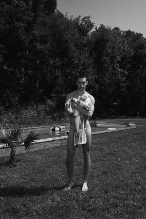Monochrome full-length portrait of a man standing outdoors by a swimming pool holding a striped towel