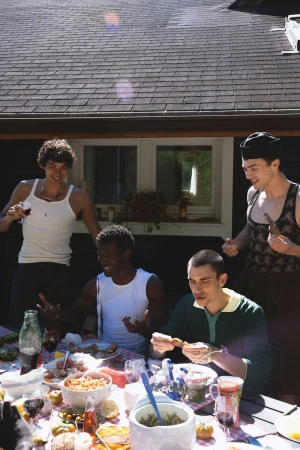 Four young men dining outdoors at a table laden with food and wine under a sunny roof overhang.