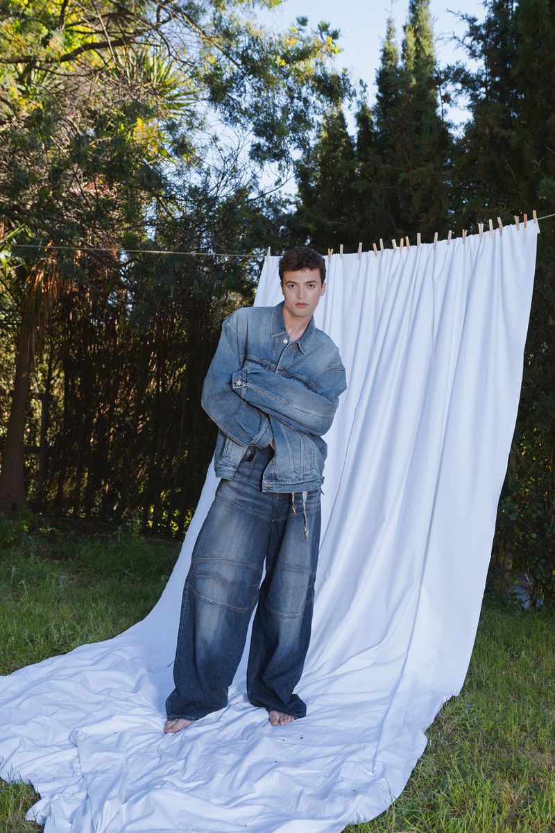 Model standing barefoot on a white cloth in a grassy garden, wearing an oversized blue denim jacket and baggy jeans.