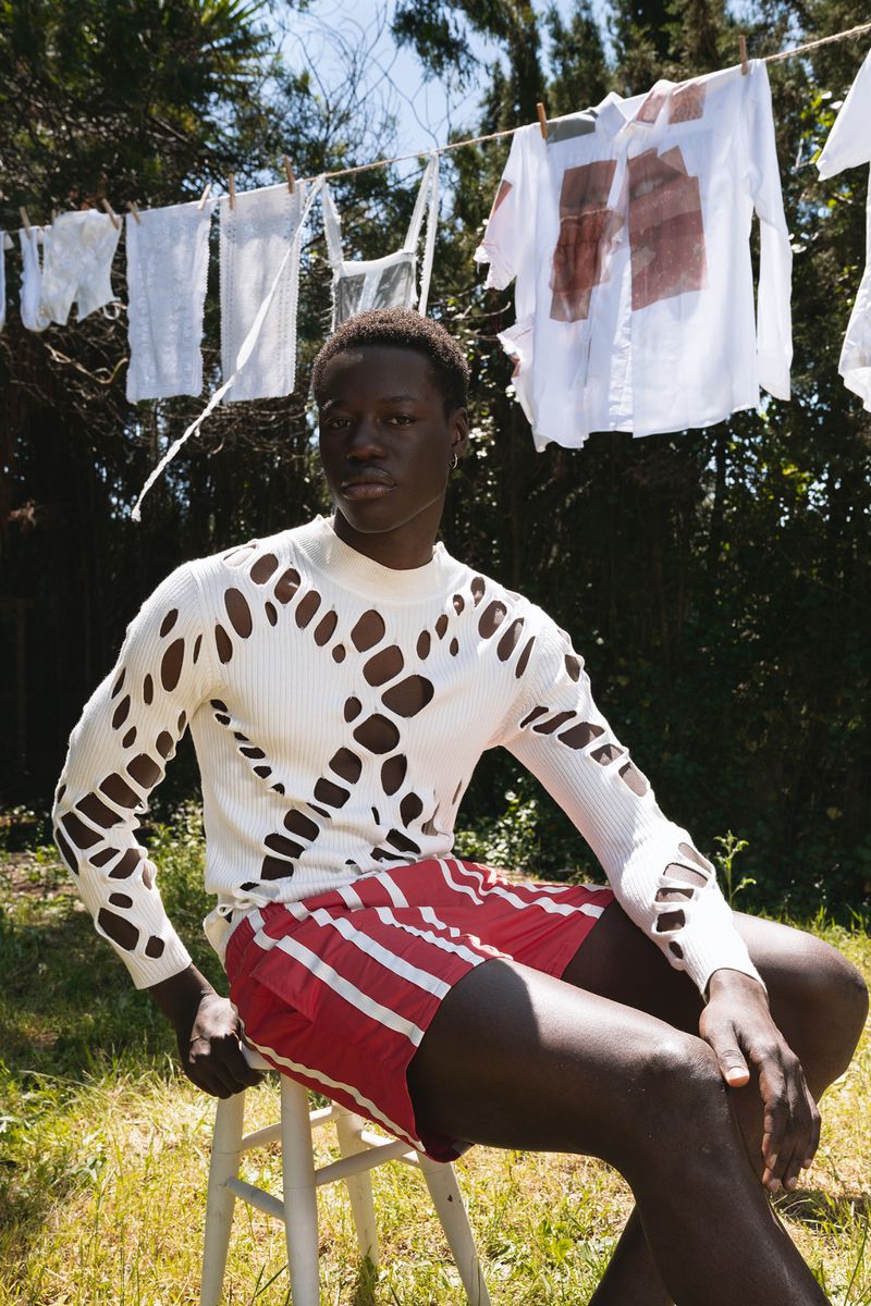 Black male model wearing a patterned white knit sweater and red shorts seated outdoors beneath a clothesline.