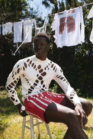 Black male model wearing a patterned white knit sweater and red shorts seated outdoors beneath a clothesline.
