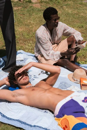 Two men relaxing on a blanket outdoors in a sunny park with books and a straw hat