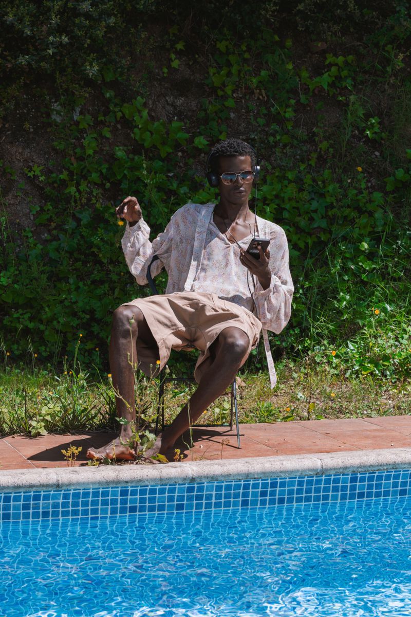 A man with sunglasses sits by a blue tiled pool wearing headphones and holding a smartphone, with a green leafy backdrop outdoors.