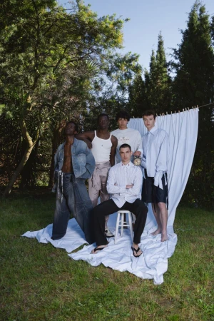 Five male models posing outdoors in an open grassy area with a white fabric backdrop draped among trees.