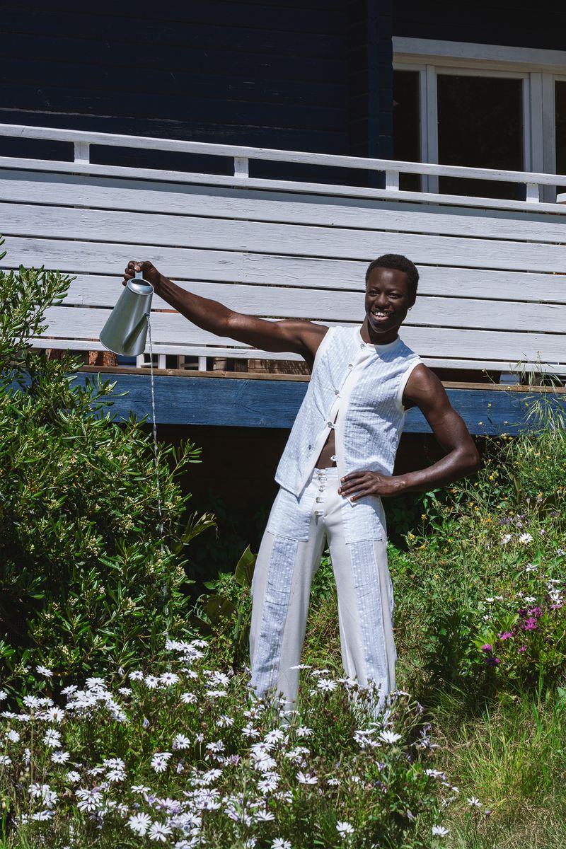 A Black man wearing a white patterned vest and matching trousers stands in a garden, watering plants with a metal watering can.