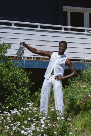 A Black man wearing a white patterned vest and matching trousers stands in a garden, watering plants with a metal watering can.
