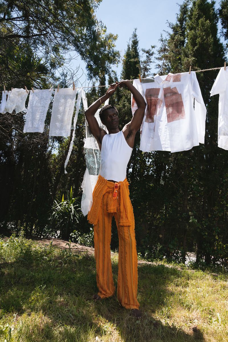 Man wearing a white tank top and orange trousers standing outdoors beneath laundry hanging on a clothesline