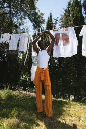 Man wearing a white tank top and orange trousers standing outdoors beneath laundry hanging on a clothesline