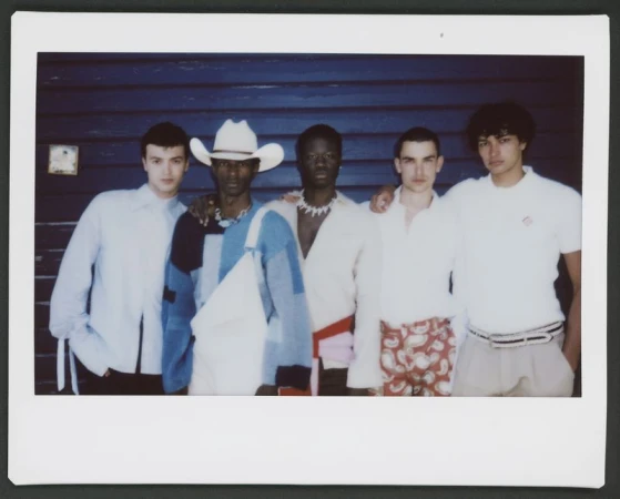 Polaroid photograph of five male models posing in front of a dark blue wooden slat wall.