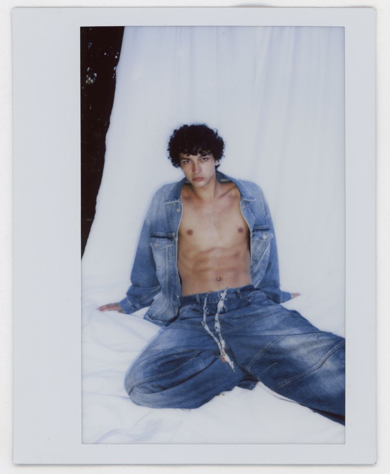 Young man with dark curly hair wearing an open denim jacket and blue denim pants seated against a white backdrop.