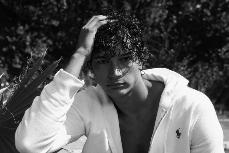 Black and white portrait of a young man with dark curly hair wearing a white hoodie, touching his hair outdoors.