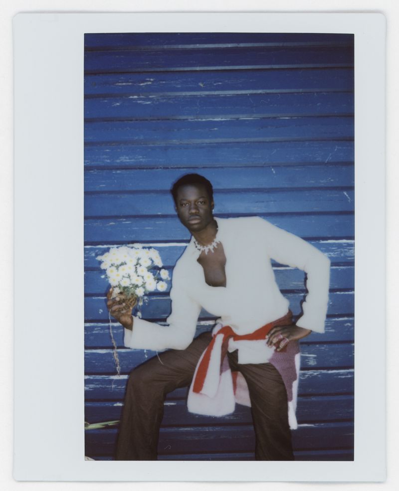 Polaroid style portrait of a Black male model wearing a white top and holding white flowers against a blue wooden wall