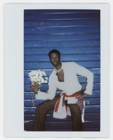 Polaroid style portrait of a Black male model wearing a white top and holding white flowers against a blue wooden wall