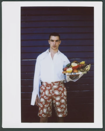 Man wearing a white shirt and patterned shorts holding a fruit platter against a dark wood wall