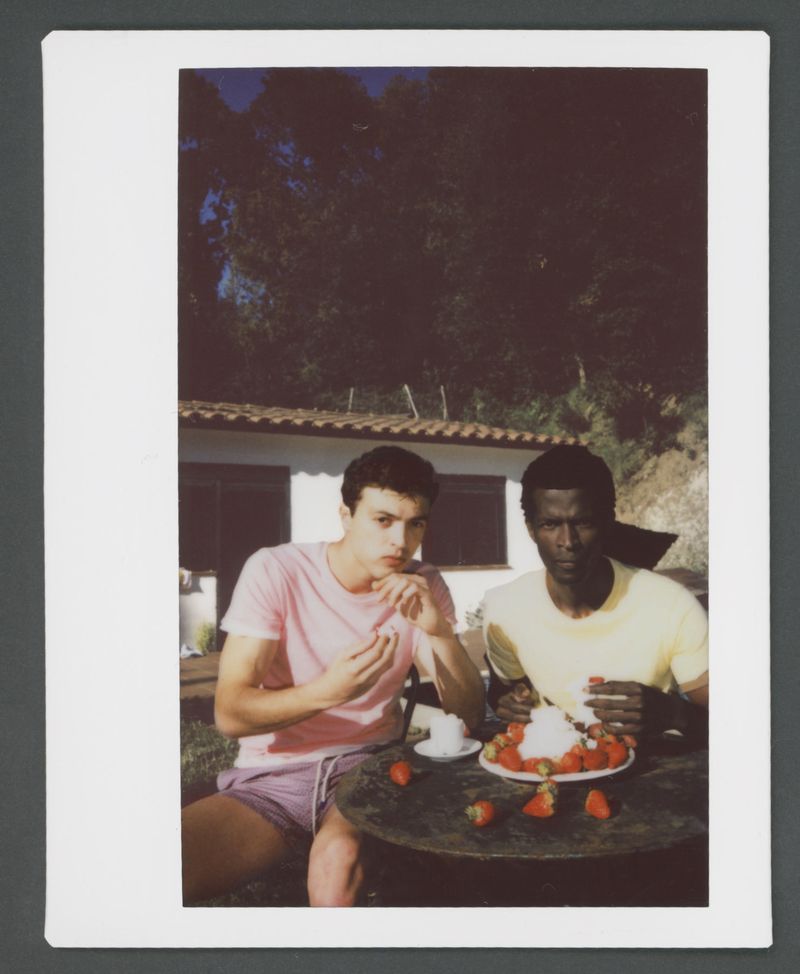 Polaroid photograph of two young men sitting outdoors at a table eating strawberries near a white building.