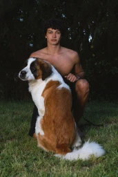 A shirtless male model kneeling outdoors on grass beside a large Saint Bernard dog against a dark foliage background.