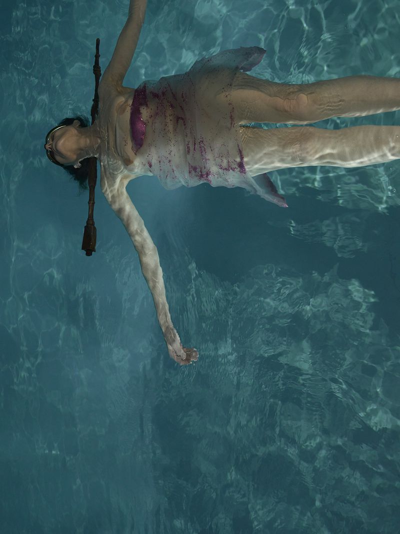 Woman in light dress floats underwater in a pool with a decorative wand. The light creates a rippling effect on the pool floor.