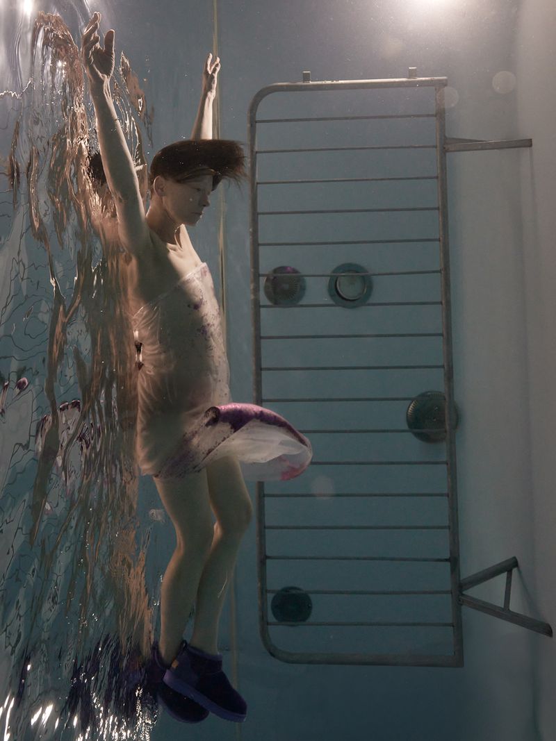 Woman submerged underwater with arms raised, wearing a wrapped translucent garment and dark shoes near a metal wall fixture.