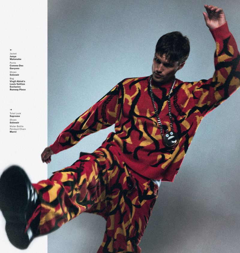 Model wearing a red and yellow patterned sweater and pants kicking upward against a grey background