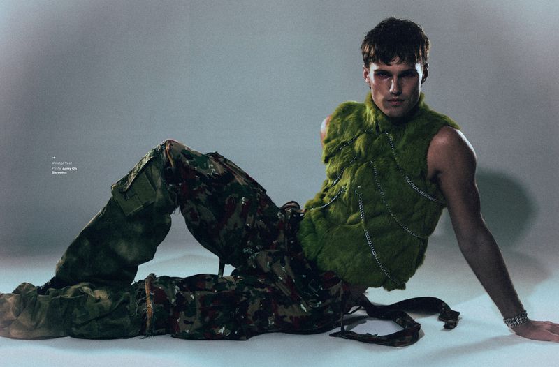 Model Nick Truelove seated wearing a green textured vest and camouflage cargo pants against a grey studio backdrop