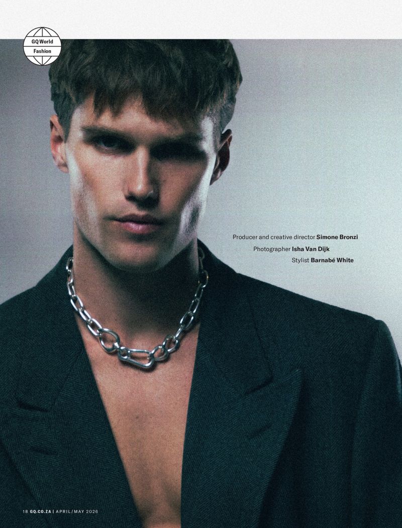 Portrait of a male model wearing a dark blazer and chunky silver chain necklace against a light grey background.