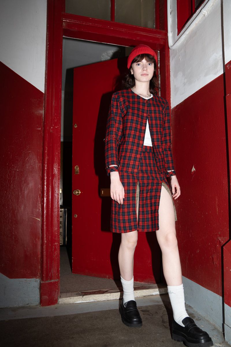 Young woman in a red tartan suit and beanie standing in an open doorway with red walls