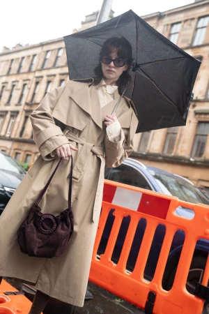 Model wearing a beige trench coat and dark sunglasses holding an umbrella and dark handbag on a city street