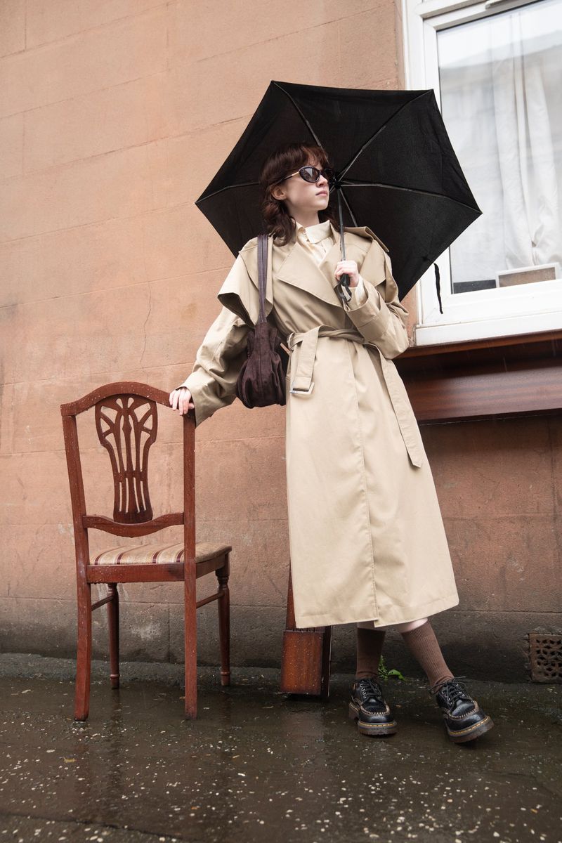 Woman in beige trench coat holding a black umbrella, leaning against a wooden chair on a wet pavement.