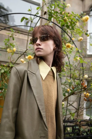 Model wearing dark sunglasses, a beige blazer, and a yellow collared shirt outdoors near yellow roses.